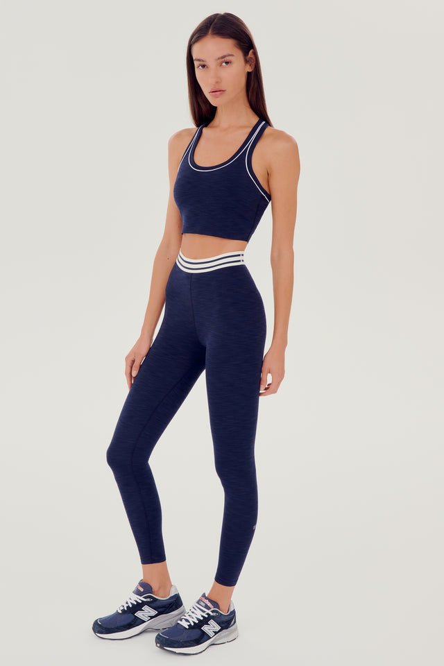 Full side view of girl wearing a dark blue ribbed bra that stops at the ribs with thin white line along arms and collar with blue leggings with white and blue waistband and blue shoes 