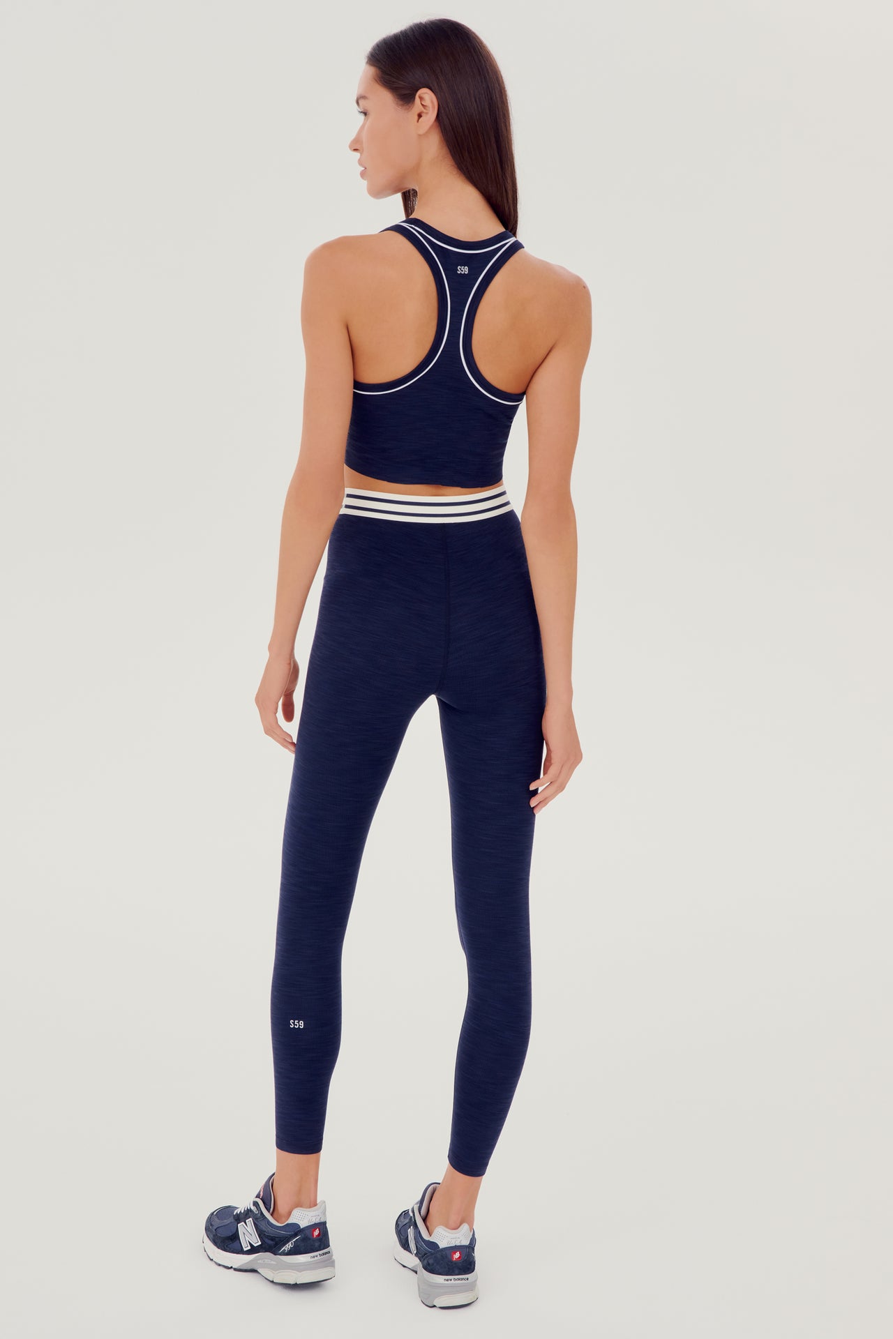 Full back view of girl wearing a dark blue ribbed bra that stops at the ribs with thin white line along arms and collar with blue leggings with white and blue waistband and blue shoes