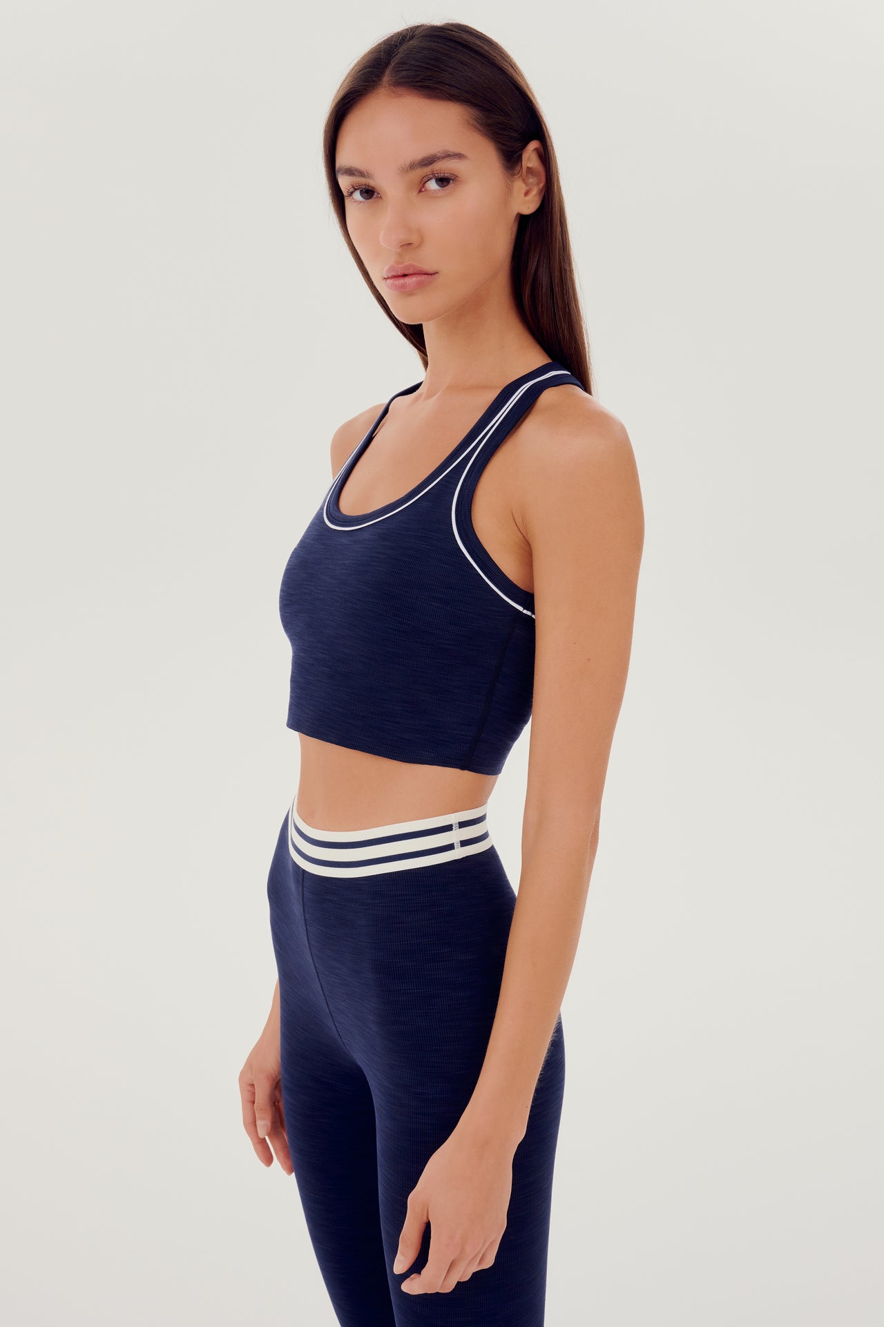 Side view of girl wearing a dark blue ribbed bra that stops at the ribs with thin white line along arms and collar with blue leggings with white and blue waistband 