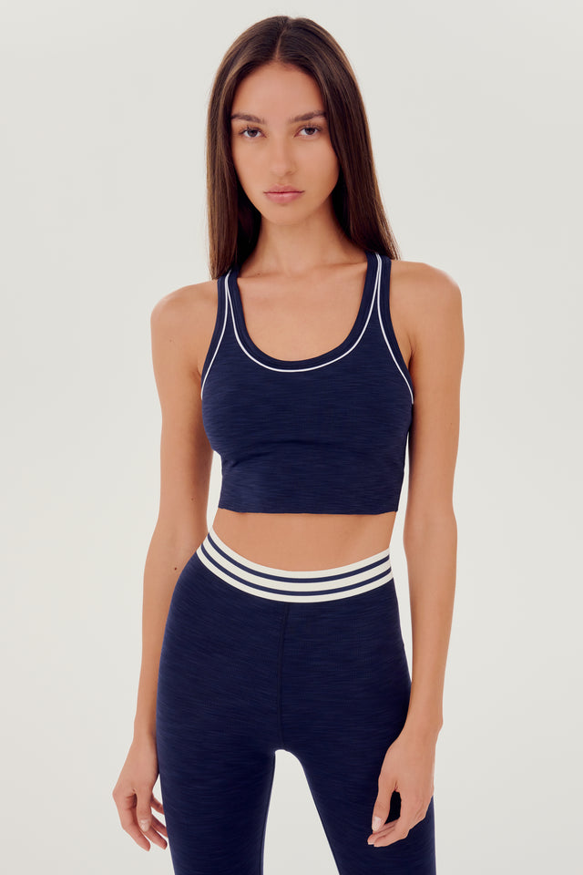 Front view of girl wearing a dark blue ribbed bra that stops at the ribs with thin white line along arms and collar with blue leggings with white and blue waistband 