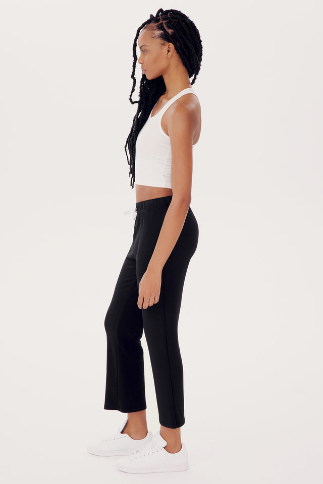 A woman with braided hair, wearing a white tank top, SPLITS59 black flared trousers, and white sneakers, standing sideways against a white background.