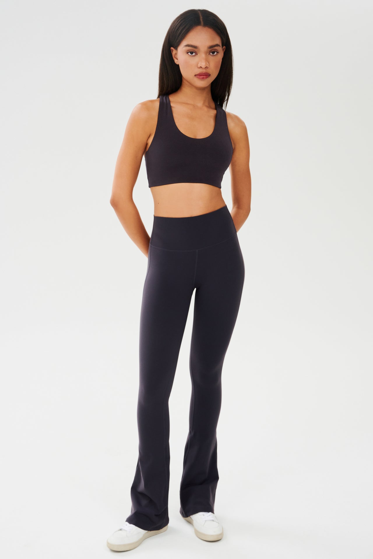 Full front view of woman with straight dark hair wearing dark gray with dark blue tone bra and dark gray with dark blue tone high waist below ankle length legging with wide flared bottoms paired with white shoes
