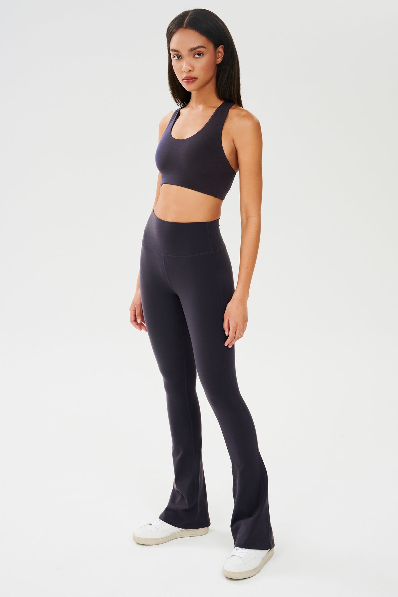 Full front side view of woman with straight dark hair wearing dark gray with dark blue tone bra and dark gray with dark blue tone leggings paired with white shoes