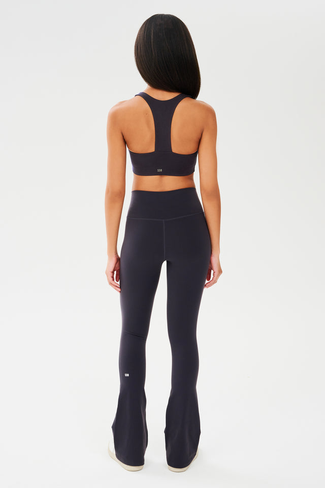 Full back view of woman with straight dark hair wearing dark gray with dark blue tone racerback bra with white S59 logo and dark gray with dark blue tone high waist below ankle length legging with wide flared bottoms and white S59 logo on bottom left calf. Paired with white shoes