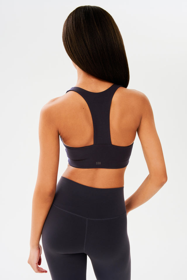 Back view of woman with straight dark hair wearing dark gray with dark blue tone bra with racerback with light grey S59 logo and dark gray with dark blue tone leggings