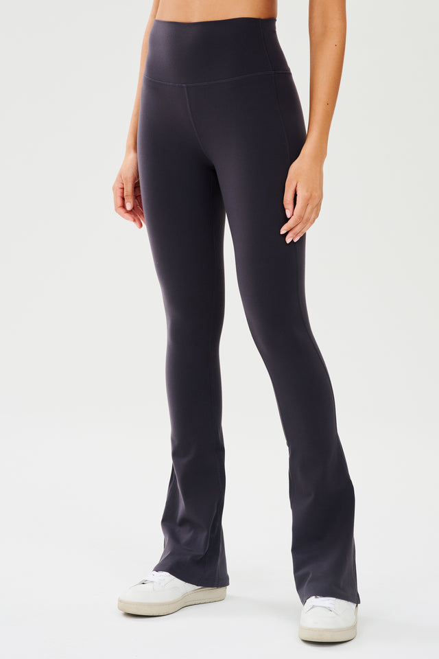 Front view of woman wearing dark gray with dark blue tone high waist below ankle length legging with wide flared bottoms paired with white shoes