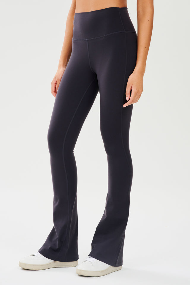 Front side view of woman wearing dark gray with dark blue tone high waist below ankle length legging with wide flared bottoms paired with white shoes