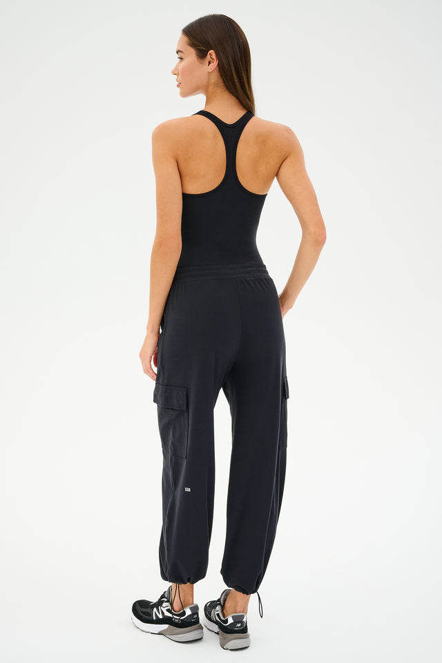 Dressed in SPLITS59's Nico Airweight Cargo Pant - Black and a black tank top, a person stands facing away from the camera in sleek black sneakers.