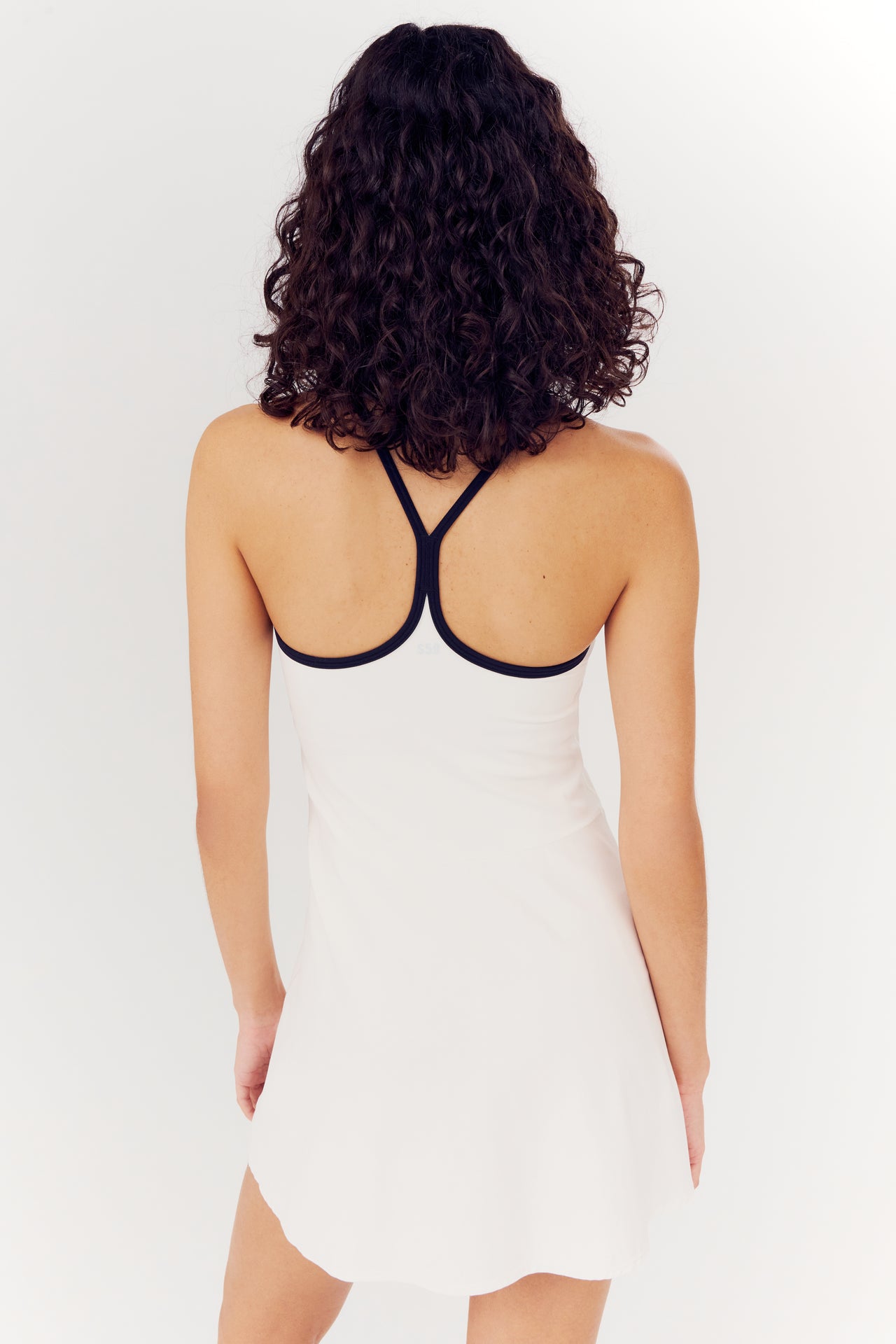 Rear view of a woman with curly hair wearing a SPLITS59 Simona Airweight Tank Dress - White/Indigo with a black spandex strap design.