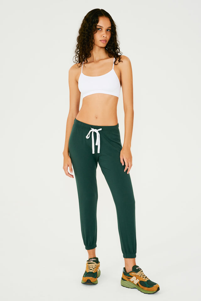 Full front view of woman with dark wavy hair wearing dark green sweatpant jogger with white drawstring and white bra with spaghetti straps paired with dark green, orange and light green shoes