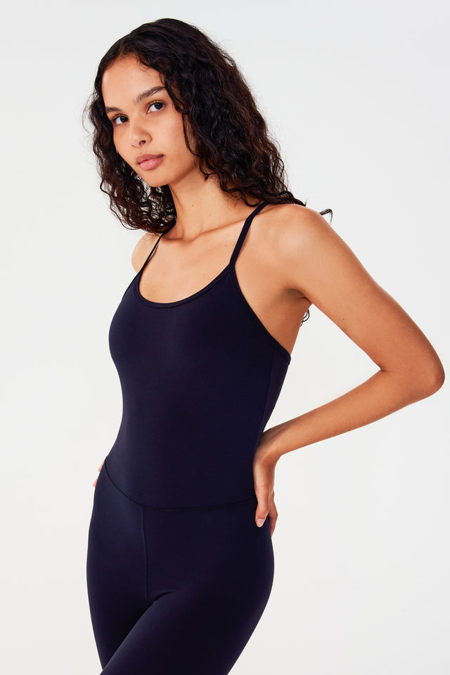 Front side view of woman with dark wavy hair wearing a dark blue one piece jumper with spaghetti straps