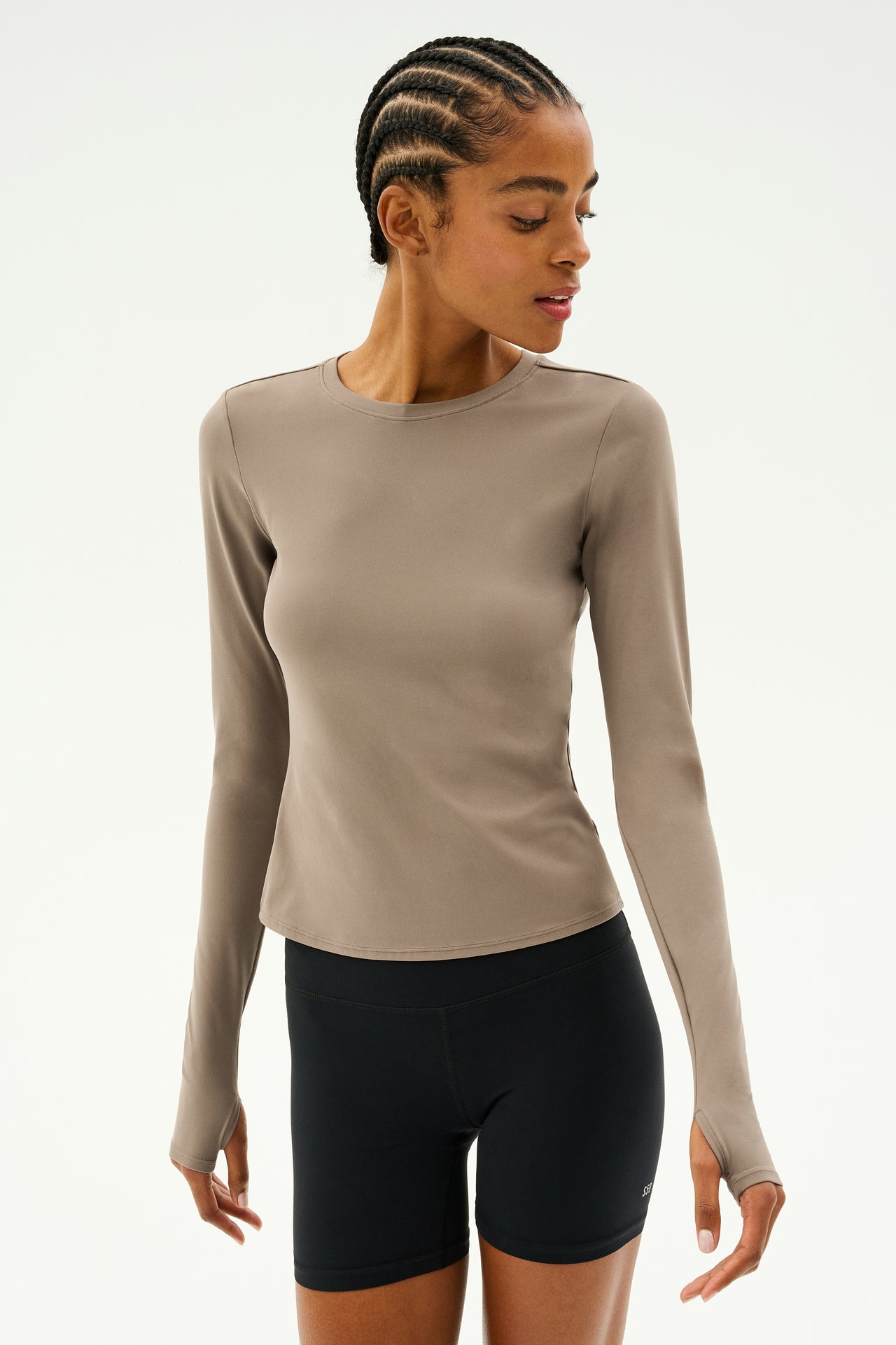 Dressed in a Lentil Airweight Lite Waist Length long-sleeve from SPLITS59 and black shorts, a person with braided hair stands against a neutral backdrop, ideal for enjoying the quick dry comfort during workouts.