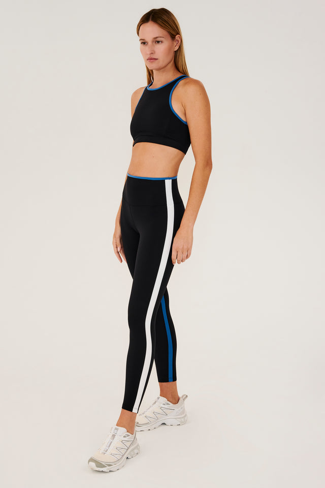 Full side view of girl wearing black sports bra with blue hem and black leggings with blue stripe around the waistband and white stripe down the outer seam and a blue strip on inseam and white shoes