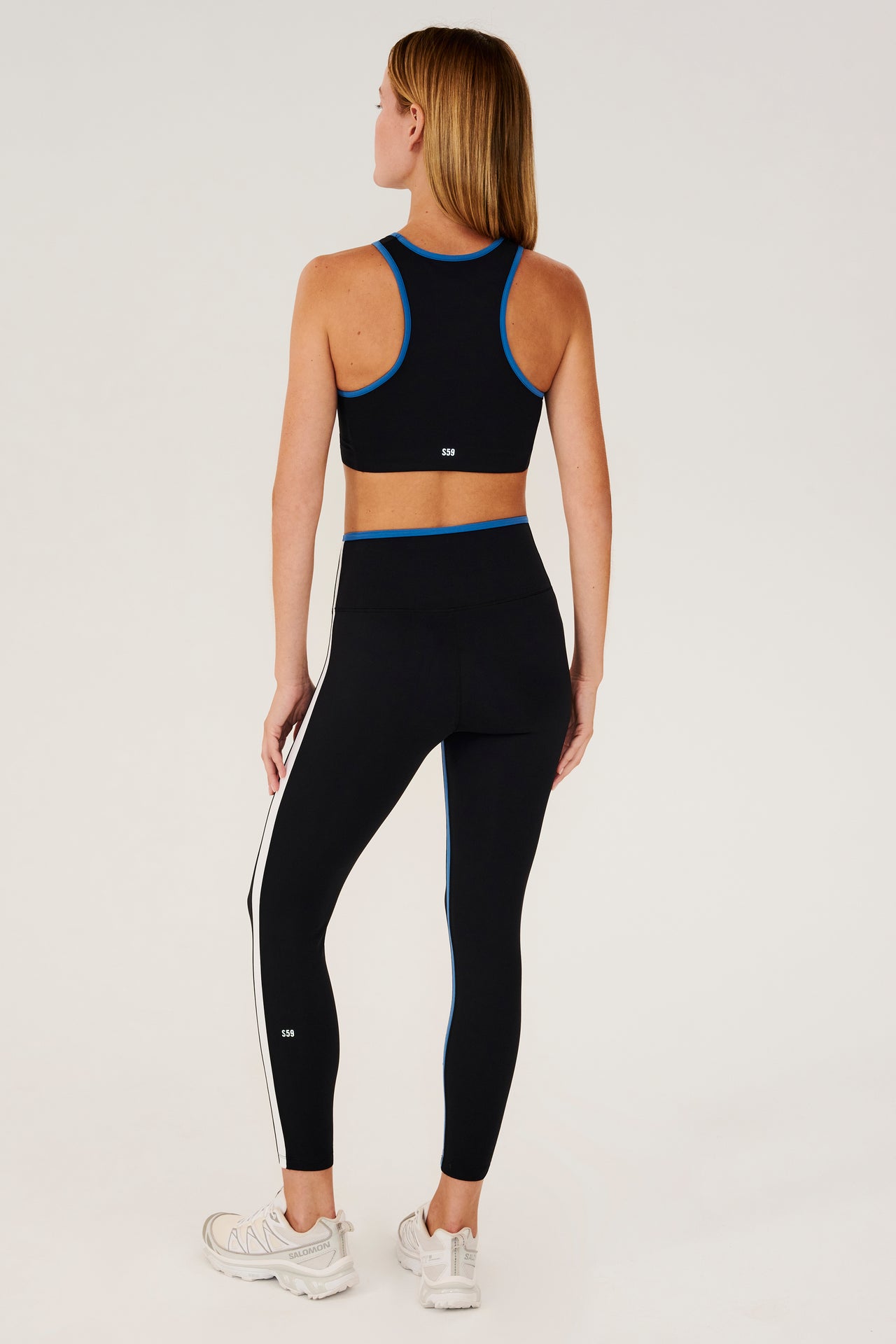 Full back view of girl wearing black sports bra with blue hem and black leggings with blue stripe around the waistband and white stripe down the outer seam and a blue strip on inseam and white shoes