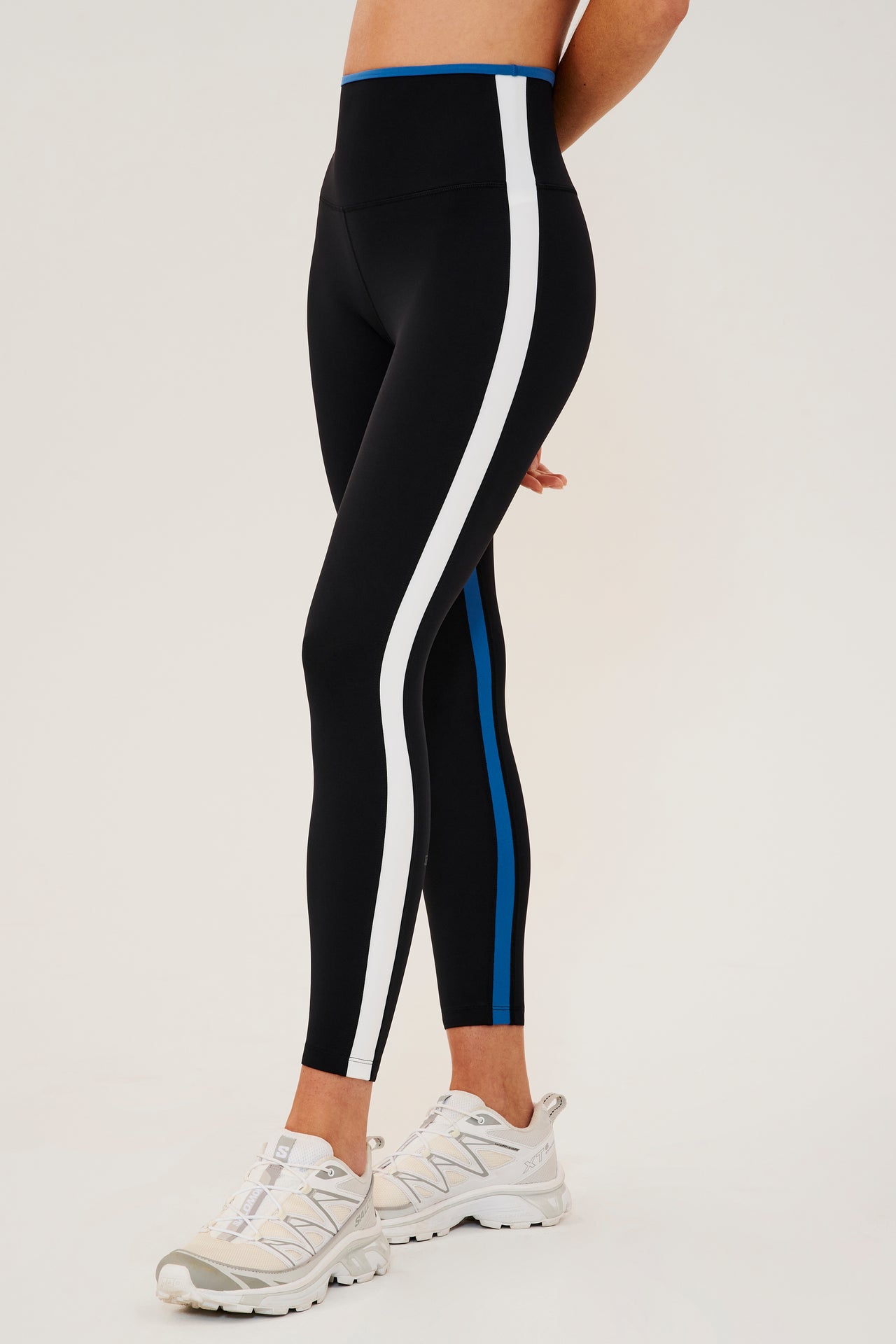 Front side view of woman wearing black leggings with bright blue and white side stripes with bright blue waistband. Paired with white shoes.