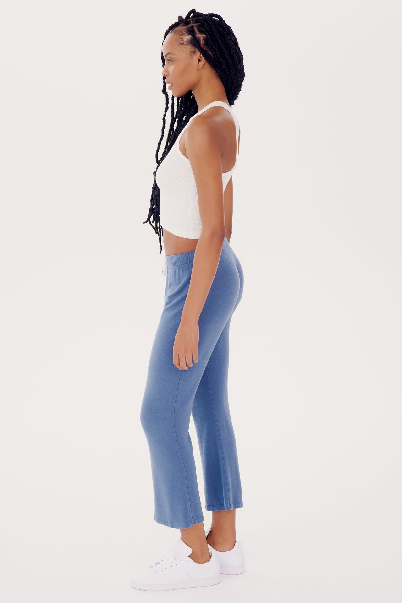 A woman in a white tank top and SPLITS59 Brooks Fleece Cropped Flare - Steel Blue pants stands in profile on a white background, wearing white sneakers.