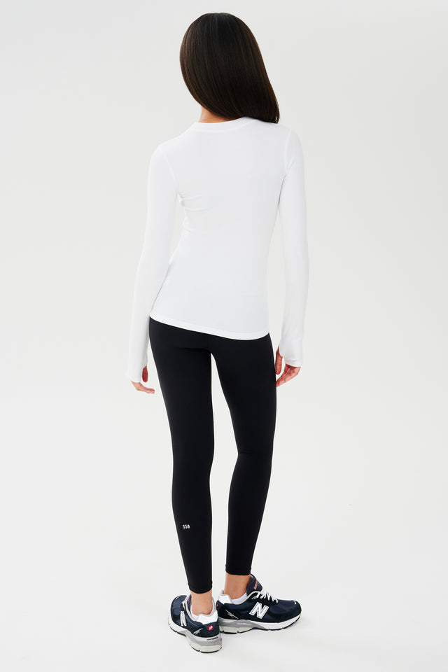 The back view of a woman wearing a SPLITS59 Louise Rib Long Sleeve - White top and black leggings, ready for her yoga session.