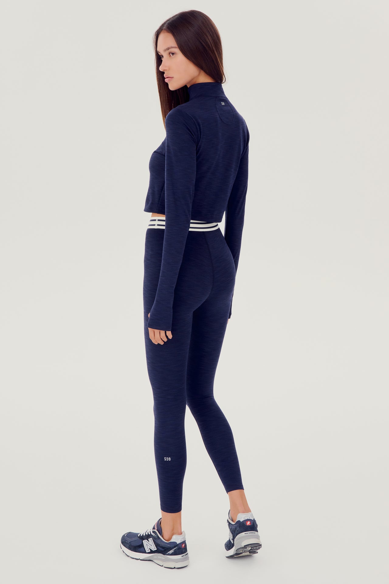 Back full view of woman with brown straight hair wearing a dark blue cropped sweatshirt with collar, and dark blue leggings with white and dark blue stripes on the waistband and wearing white and dark blue shoes