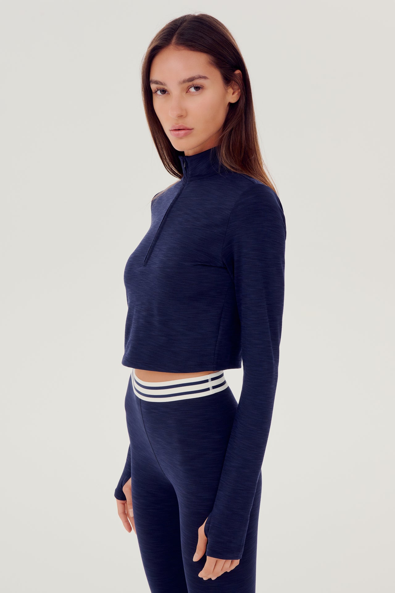 Front view of woman with brown straight hair wearing a dark blue cropped sweatshirt with collar, and dark blue leggings with white and dark blue stripes on the waistband
