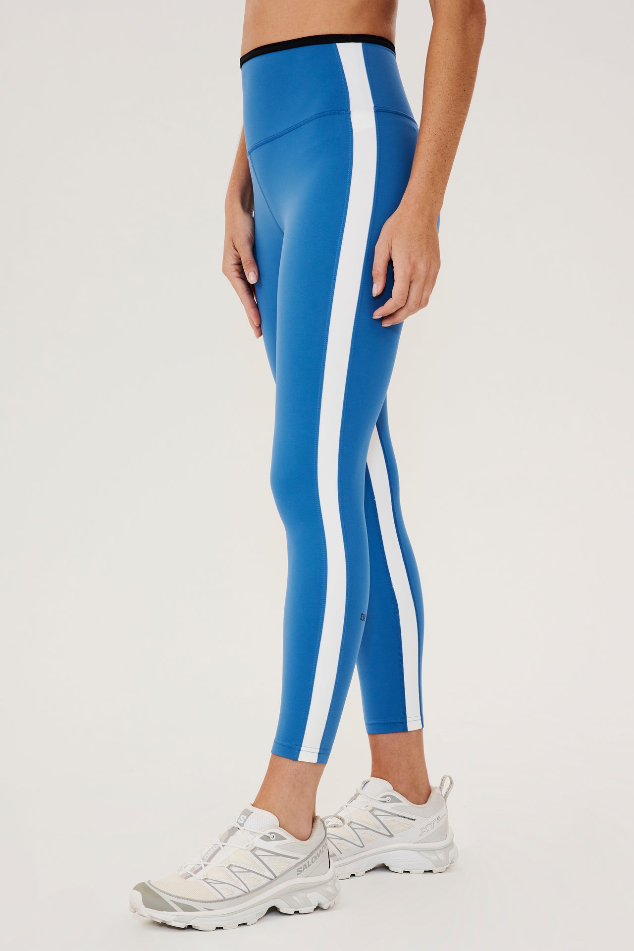 Front side view of woman wearing bright blue leggings with white side stripes and black waistband paired with white shoes
