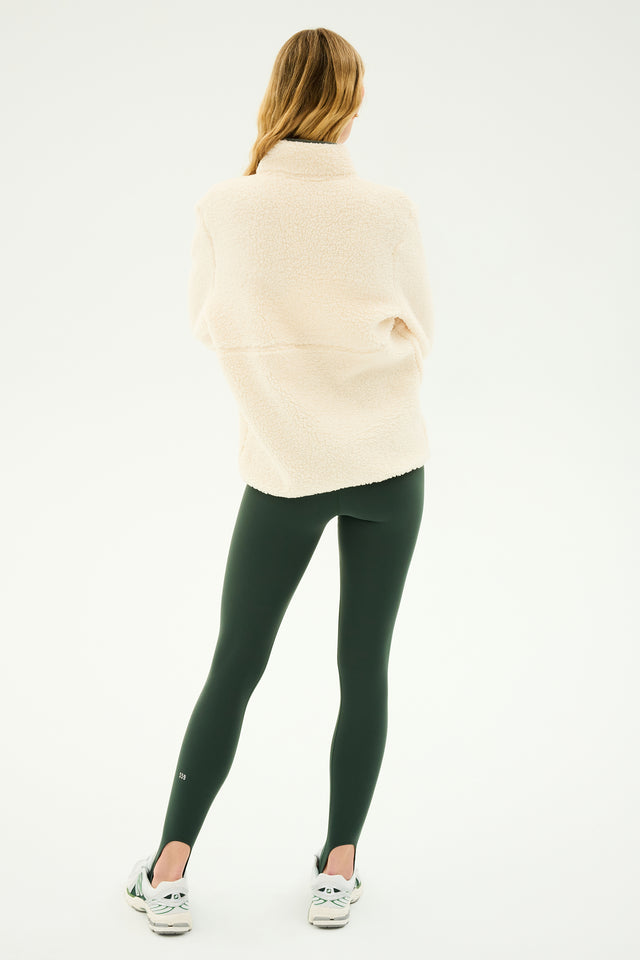 A person with long hair is standing with their back to the camera, wearing SPLITS59's Libby Sherpa Half Zip in Creme/Military along with dark green leggings and white sneakers—ready for winter workouts or perhaps some yoga practice.