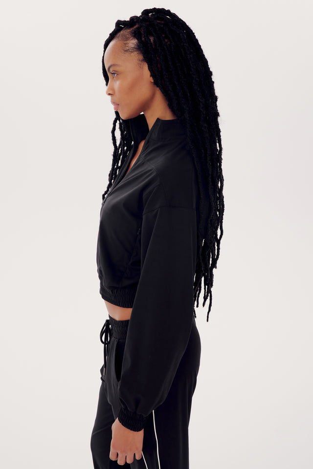 Side profile of a woman with long braids wearing a SPLITS59 Harlowe Rigor Jacket in Black and pants against a white background.