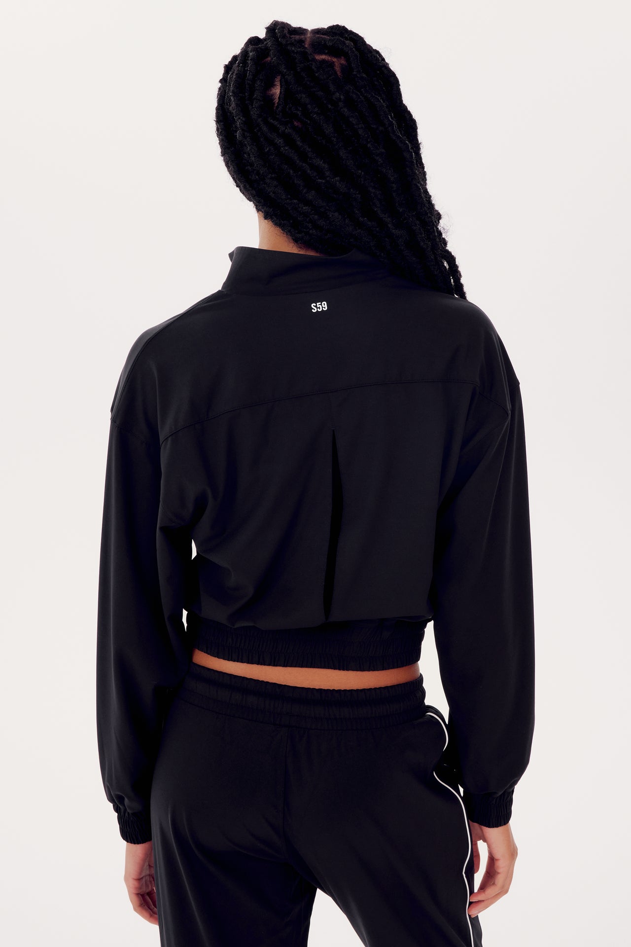 A person seen from the back wearing a SPLITS59 Harlowe Rigor Jacket in Black and black pants with a logo at the upper back. The person has long braided hair.