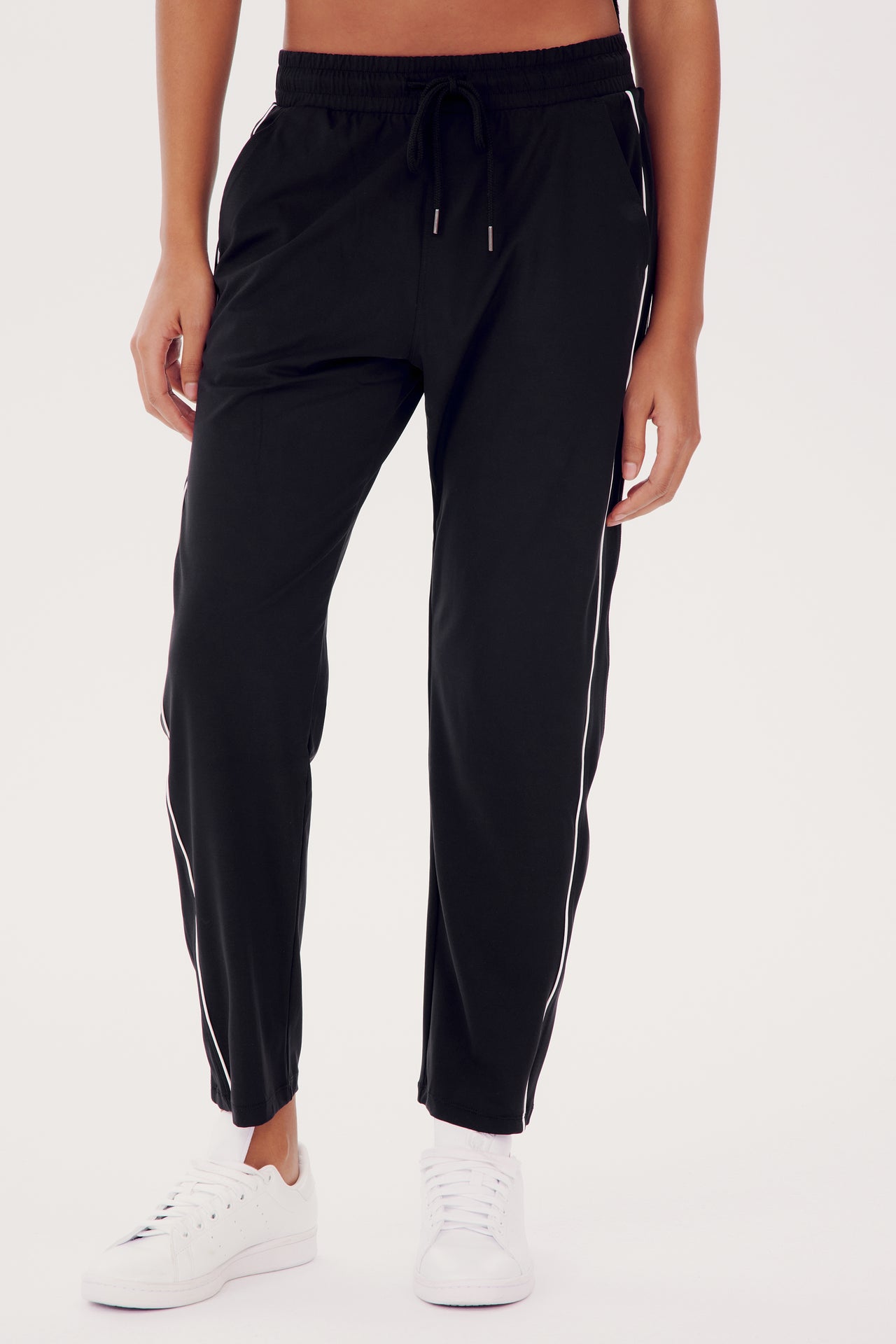 A person wearing SPLITS59 Lucy Rigor Pant W/Piping in Black with a white stripe on the sides, paired with white sneakers, stands against a plain background.