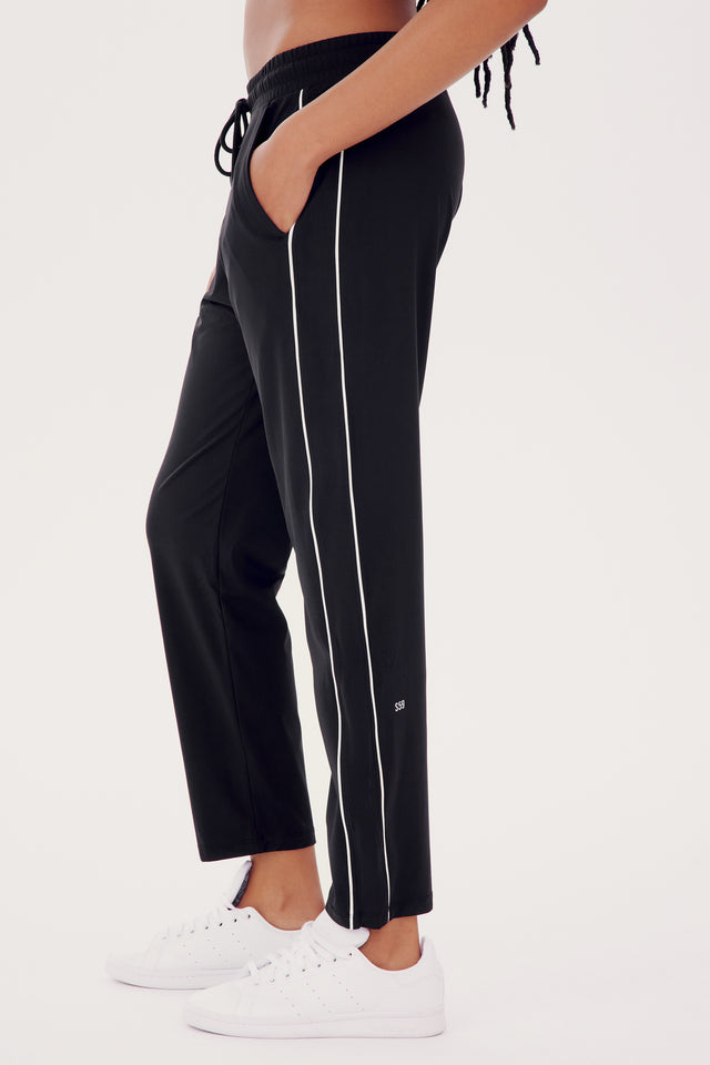 Side view of a person wearing SPLITS59 Lucy Rigor Pant W/Piping in Black with white stripes and white sneakers, standing on a plain background.