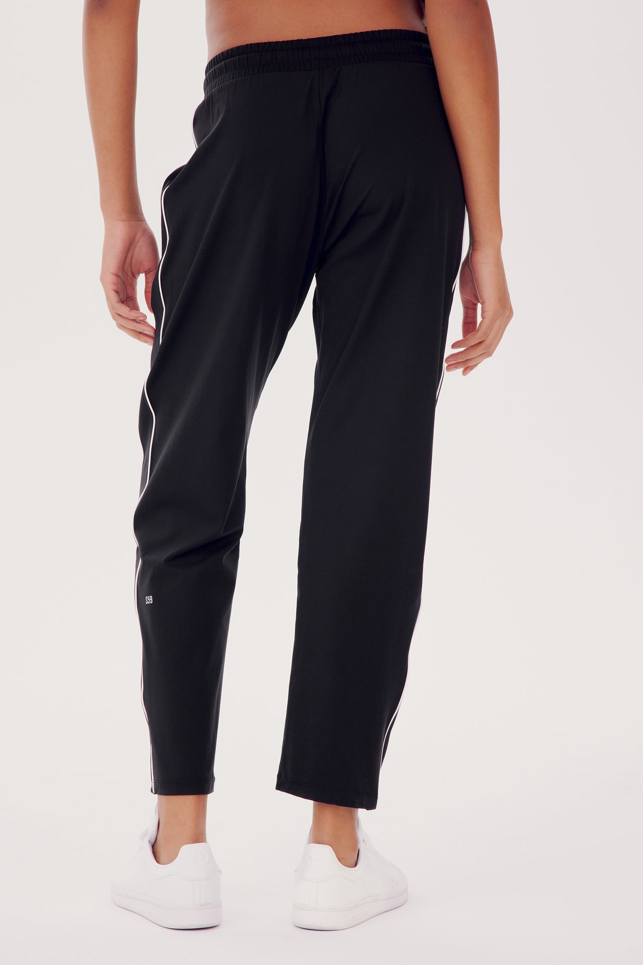 A person wearing SPLITS59 Lucy Rigor Pant W/Piping in Black with a white stripe down the side and white sneakers, standing with their back turned.
