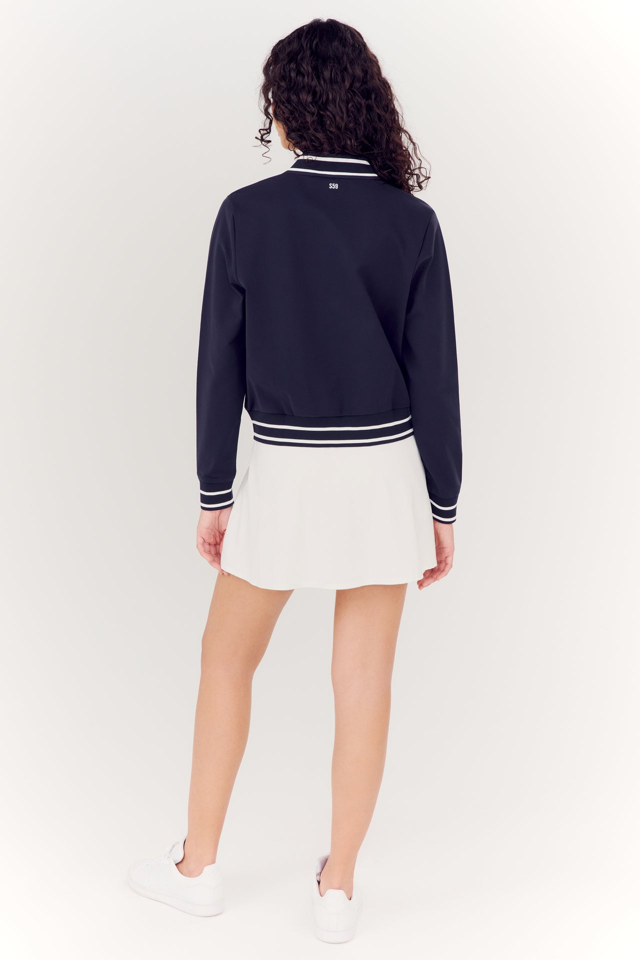 A woman seen from behind wearing a SPLITS59 Ever Supplex Jacket in Indigo and a white pleated skirt made of nylon spandex fabric with white sneakers.
