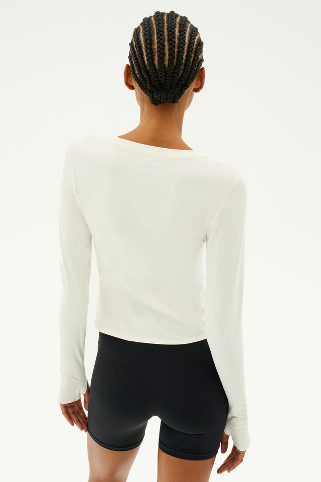 A person with braided hair is standing with their back to the camera, wearing the Airweight Lite LS Waist Length top in white from SPLITS59, paired with black quick-dry comfort shorts.