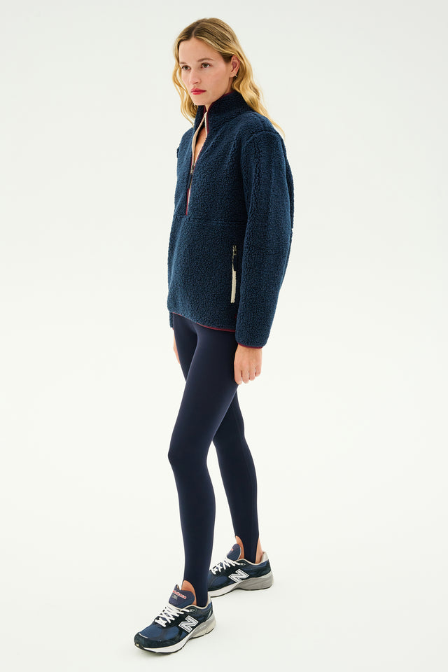 A person dressed in a SPLITS59 Libby Sherpa Half Zip in Indigo/Dark Cherry, black leggings, and navy sneakers stands against a plain white background, ready for winter workouts.