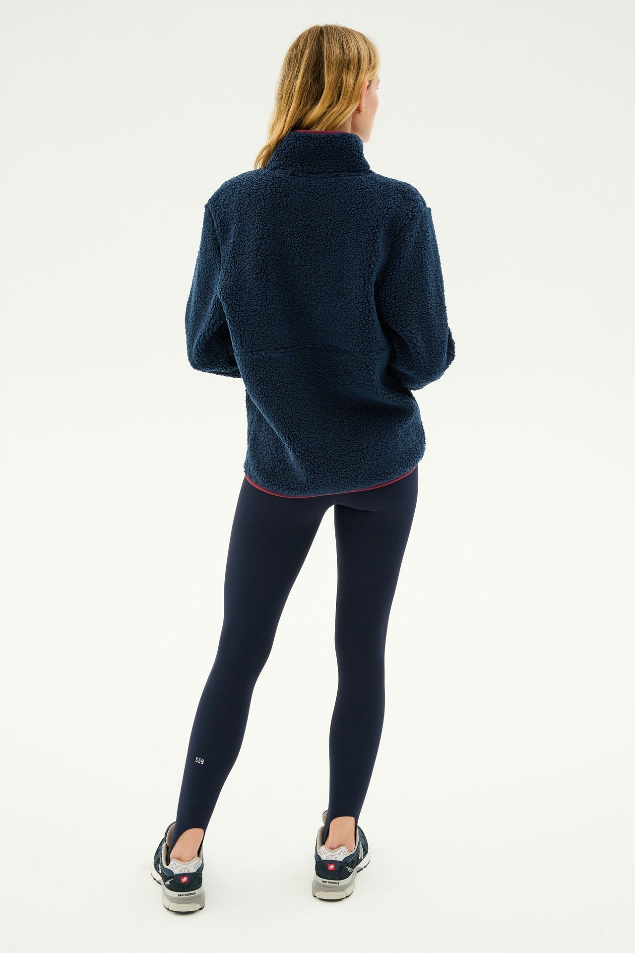 A person with long blond hair stands facing away, wearing the SPLITS59 Libby Sherpa Half Zip in Indigo/Dark Cherry, black leggings, and black shoes with white soles against a plain white background. Perfect gear for winter workouts or yoga sessions.