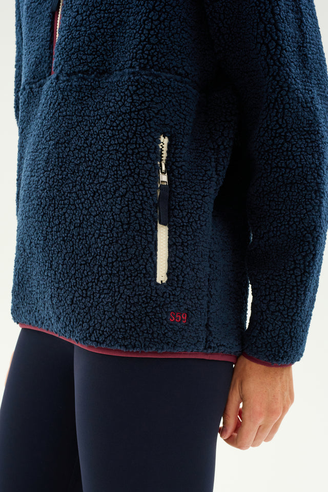 A person is wearing a SPLITS59 Libby Sherpa Half Zip in Indigo/Dark Cherry with a visible zippered pocket on the side, paired with matching navy blue pants, standing against a plain background—an ideal outfit for winter workouts.