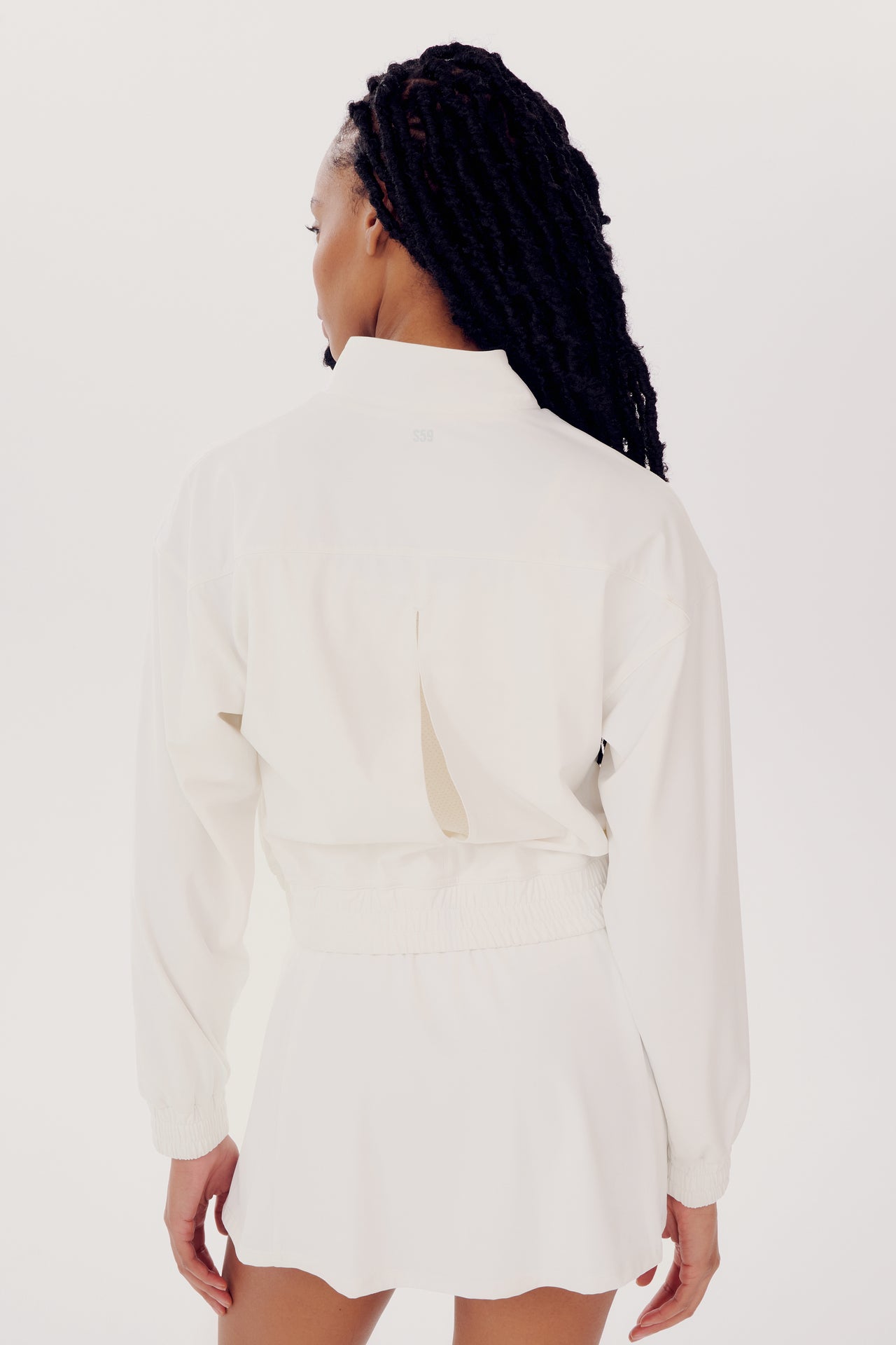 Rear view of a woman wearing a stylish white blouse and shorts, complemented by a SPLITS59 Harlowe Rigor Jacket in White, standing against a plain background. Her long braided hair cascades down her back.