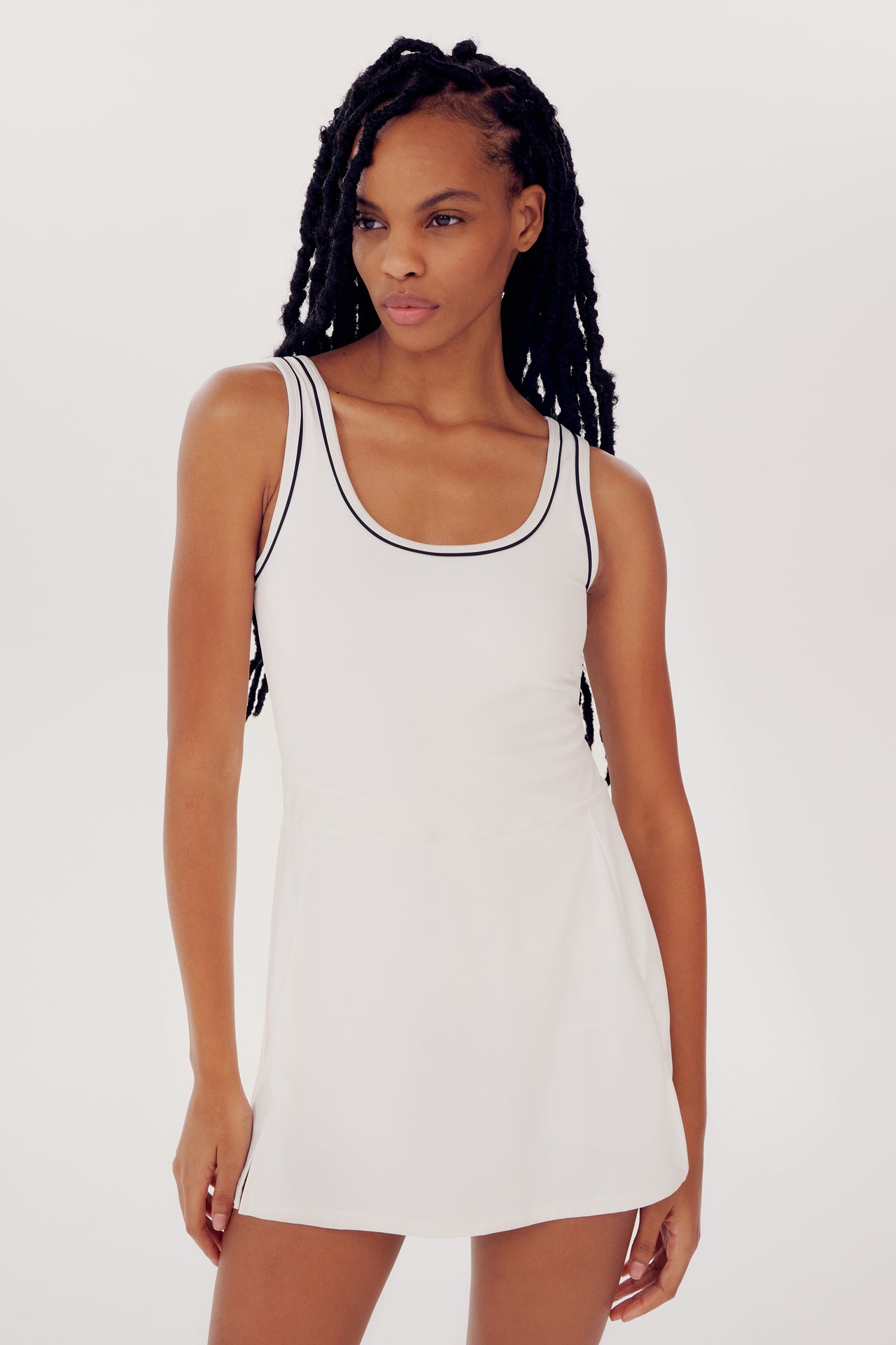 A person with long braided hair wears a SPLITS59 Martina Rigor Dress w/Piping - White, a white sleeveless design with black trim. They are standing against a plain, light-colored background.