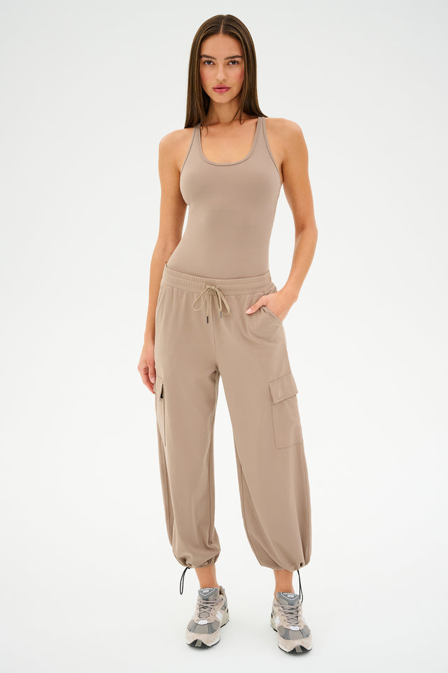 A woman stands confidently in SPLITS59's Nico Airweight Cargo Pants - Latte, a beige tank top, and sneakers. She has long hair and one hand in her pocket.