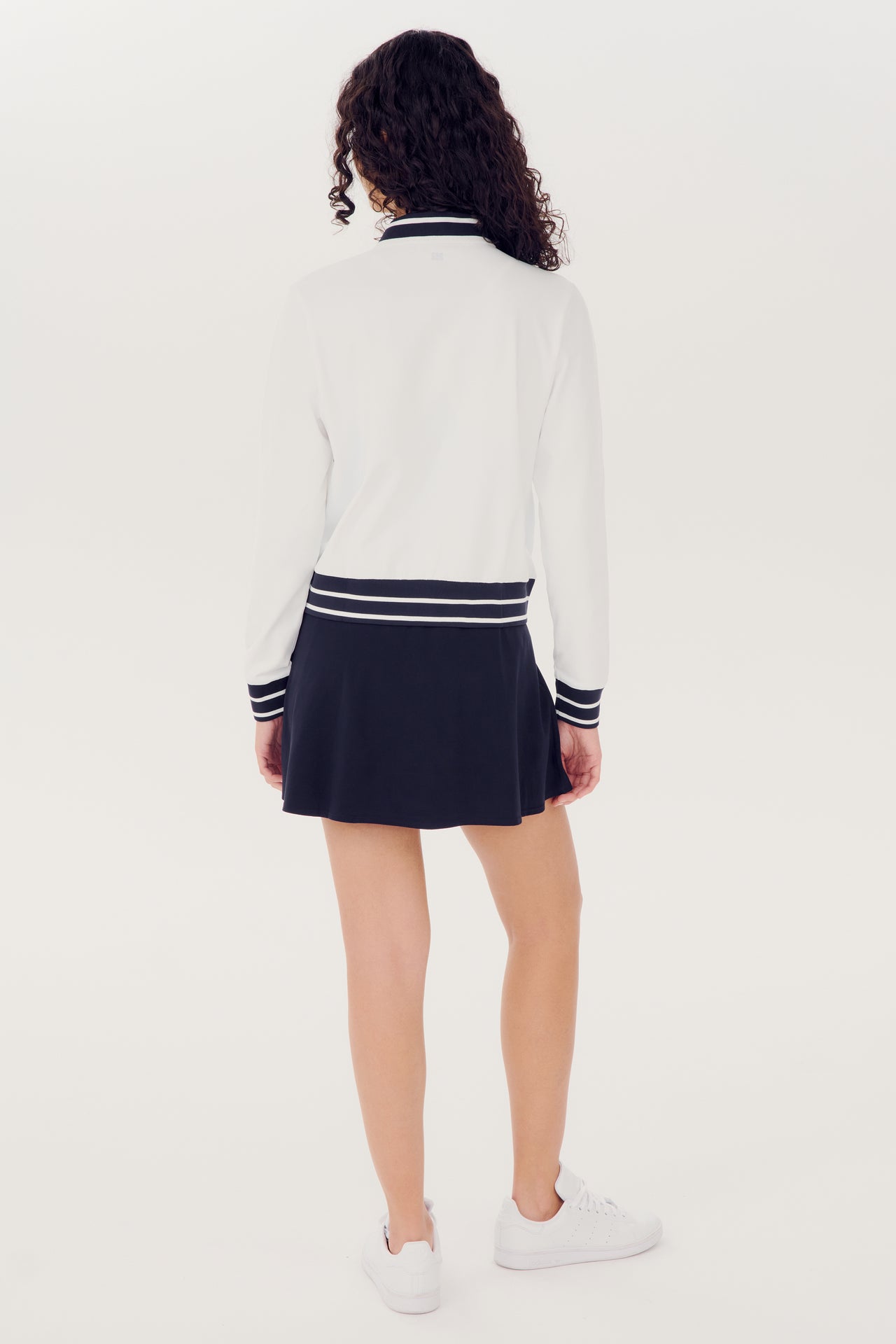 A person standing with their back to the camera wearing a SPLITS59 Ever Supplex Jacket in White with black trim and a navy skirt with white stripes, paired with white sneakers.