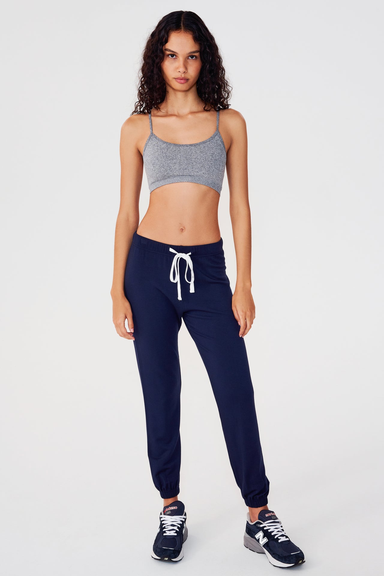Full front view of woman with dark wavy hair wearing dark blue sweatpant jogger with white drawstring and dark grey bra with spaghetti straps paired with dark blue and white shoes