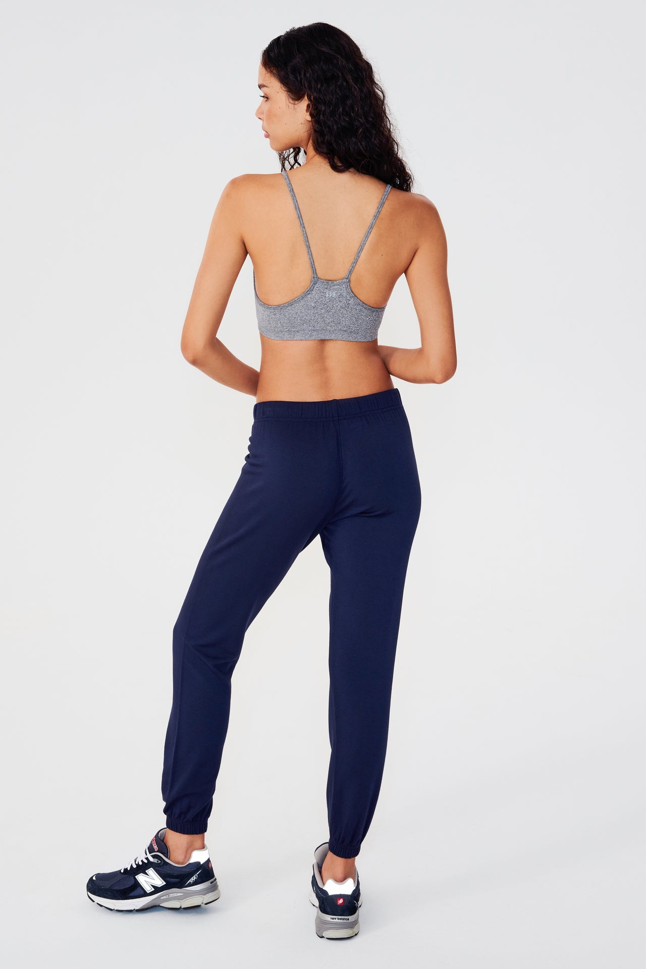Full back view of woman with dark wavy hair wearing dark blue sweatpant jogger with white drawstring and dark grey bra with racerback spaghetti straps paired with dark blue and white shoes   