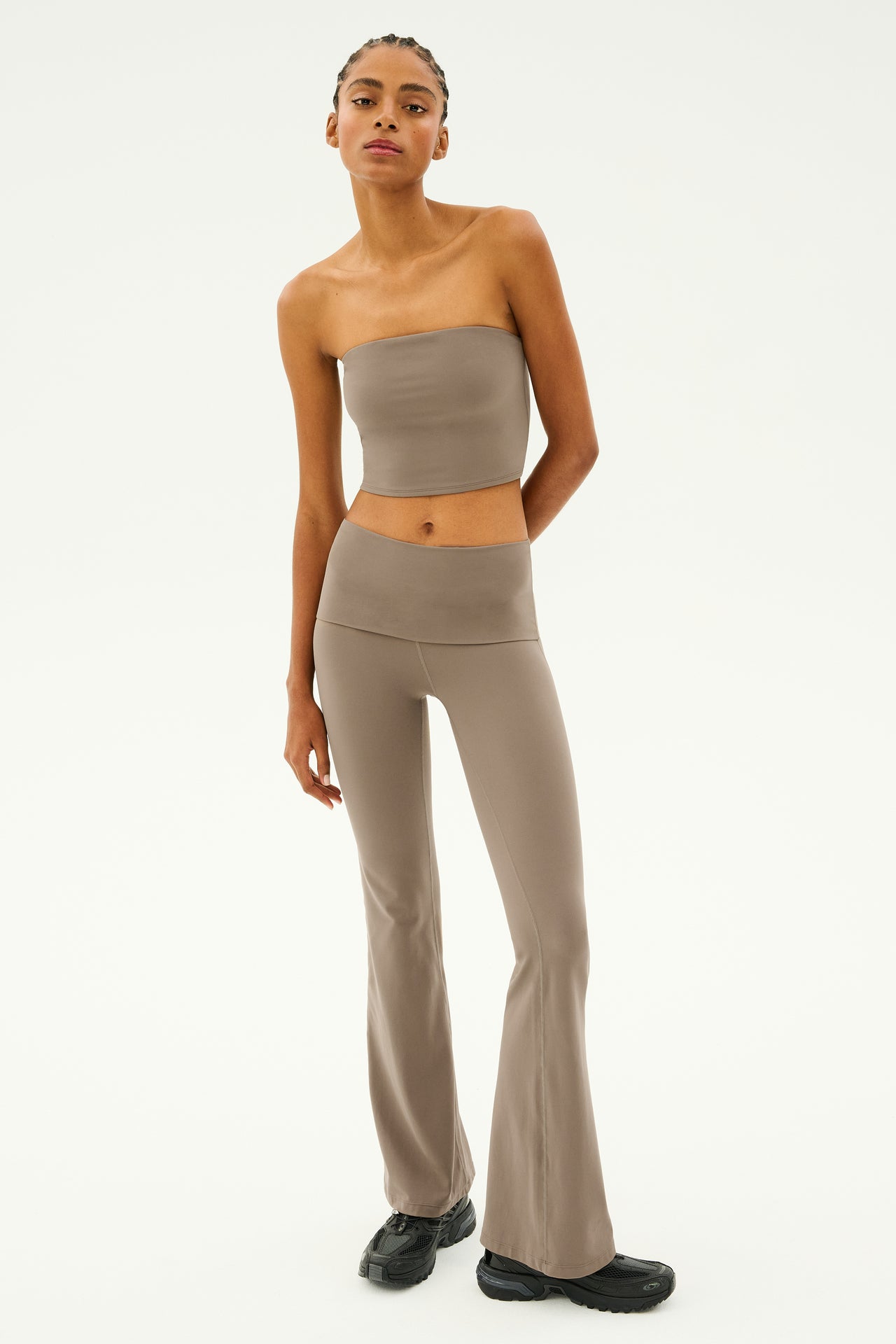 A person wearing the SPLITS59 Hazel Airweight Crop Bandeau in Lentil, a taupe bandeau-style strapless top, paired with flared pants made of stretch fabric and black shoes stands against a plain background.