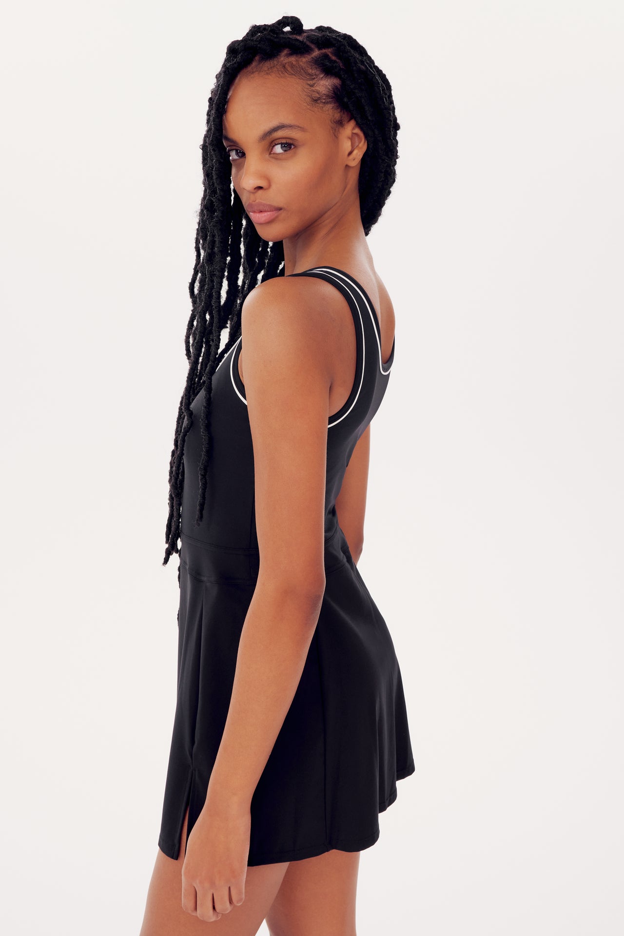 A woman with long braided hair wearing a Martina Rigor Dress W/Piping in black from SPLITS59, looking over her shoulder against a plain white background.