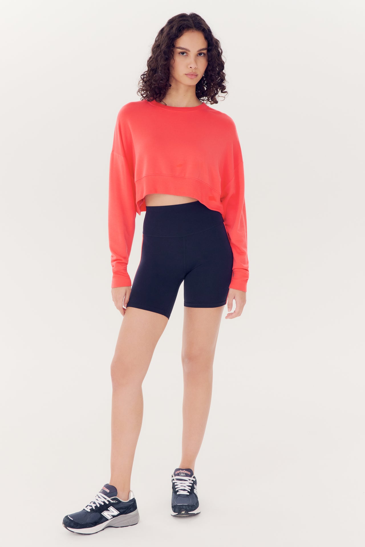 A woman stands posing against a white background, wearing a SPLITS59 Noah Fleece Crop Sweatshirt in Melon made of modal spandex blend, black shorts, and black athletic shoes.