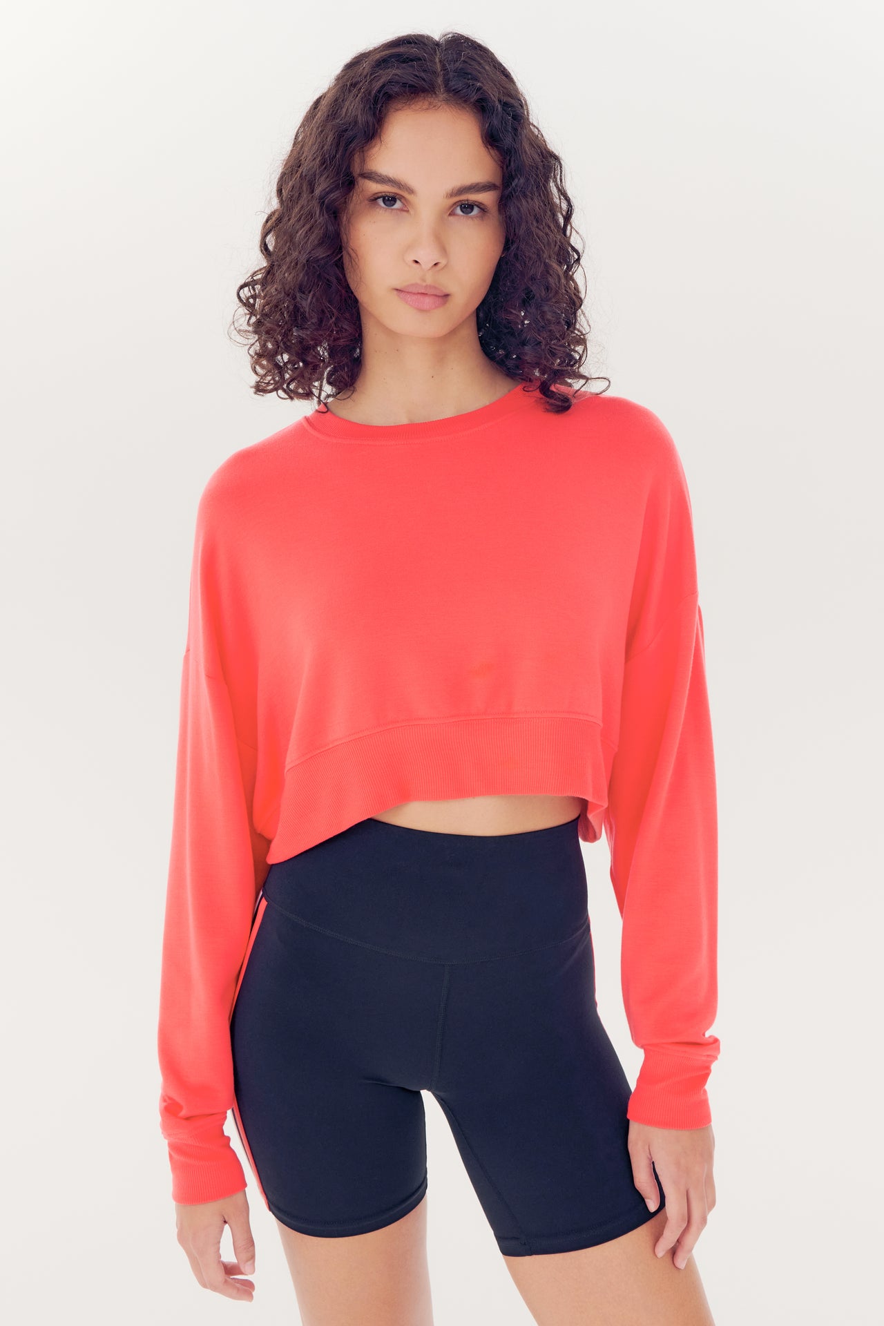 A young woman wearing a Noah Fleece Crop Sweatshirt in melon by SPLITS59, and black shorts, both made of modal spandex blend, against a white background.