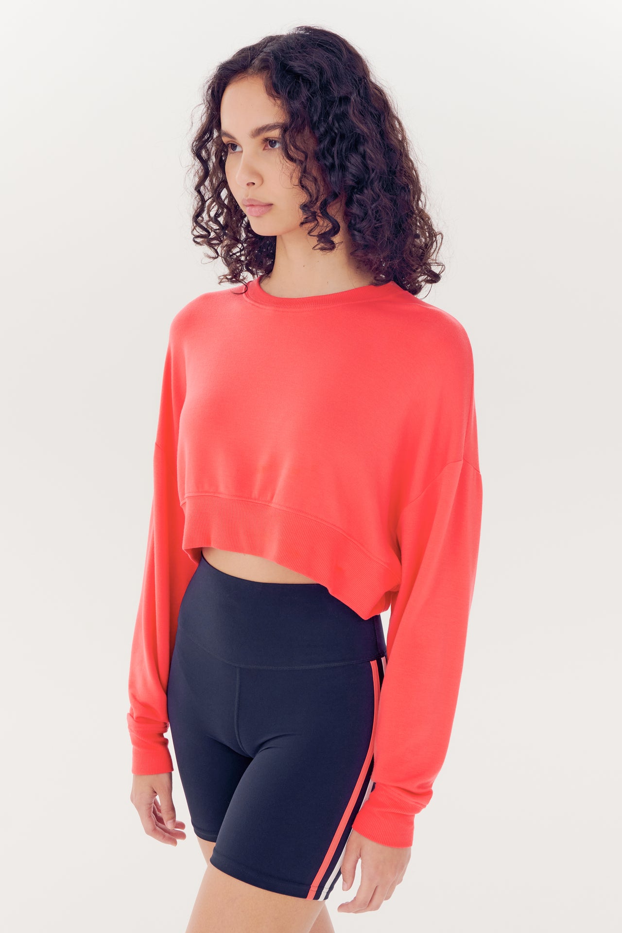 Woman in red Noah Fleece Crop Sweatshirt by SPLITS59 and black shorts made of modal spandex blend against a white background.