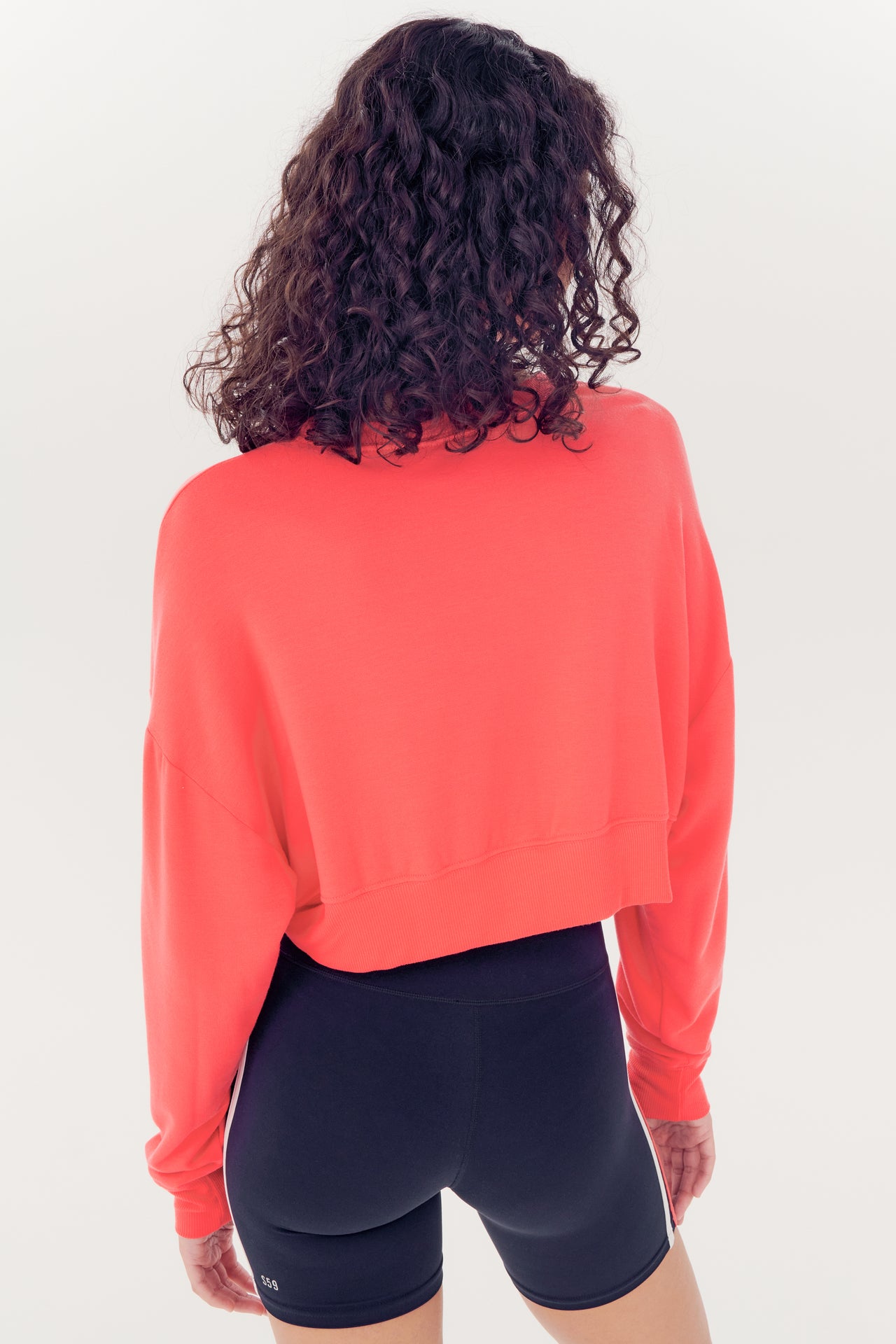 Woman standing with her back to the camera, wearing a coral red Noah Fleece Crop Sweatshirt made of modal spandex blend and black shorts by SPLITS59.