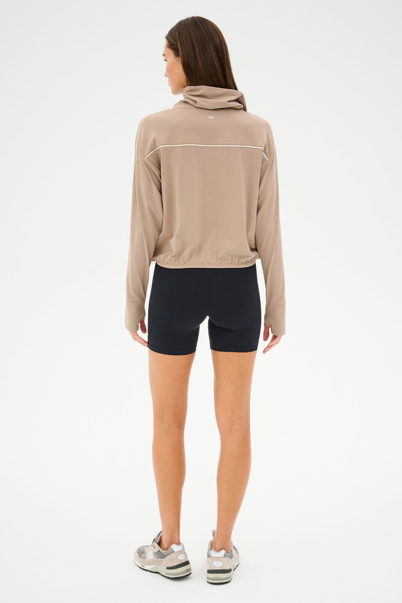 A person in the SPLITS59 Rosie Airweight Jacket - Latte, paired with black shorts and sneakers, stands facing away against a plain white background, showcasing the buttery soft fabric that hugs their frame with aerodynamic lines.