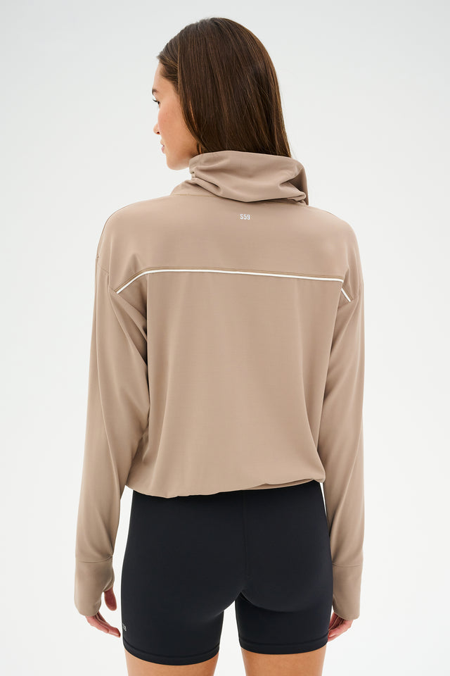 Woman in the Rosie Airweight Jacket by SPLITS59 in Latte and black shorts, facing away, showcasing buttery soft fabric ideal for hot yoga sessions.