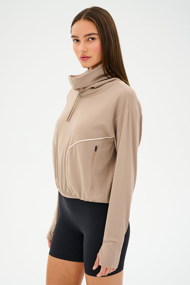 A person wearing the SPLITS59 Rosie Airweight Jacket in Latte, featuring buttery soft fabric, and black shorts stands against a plain background.